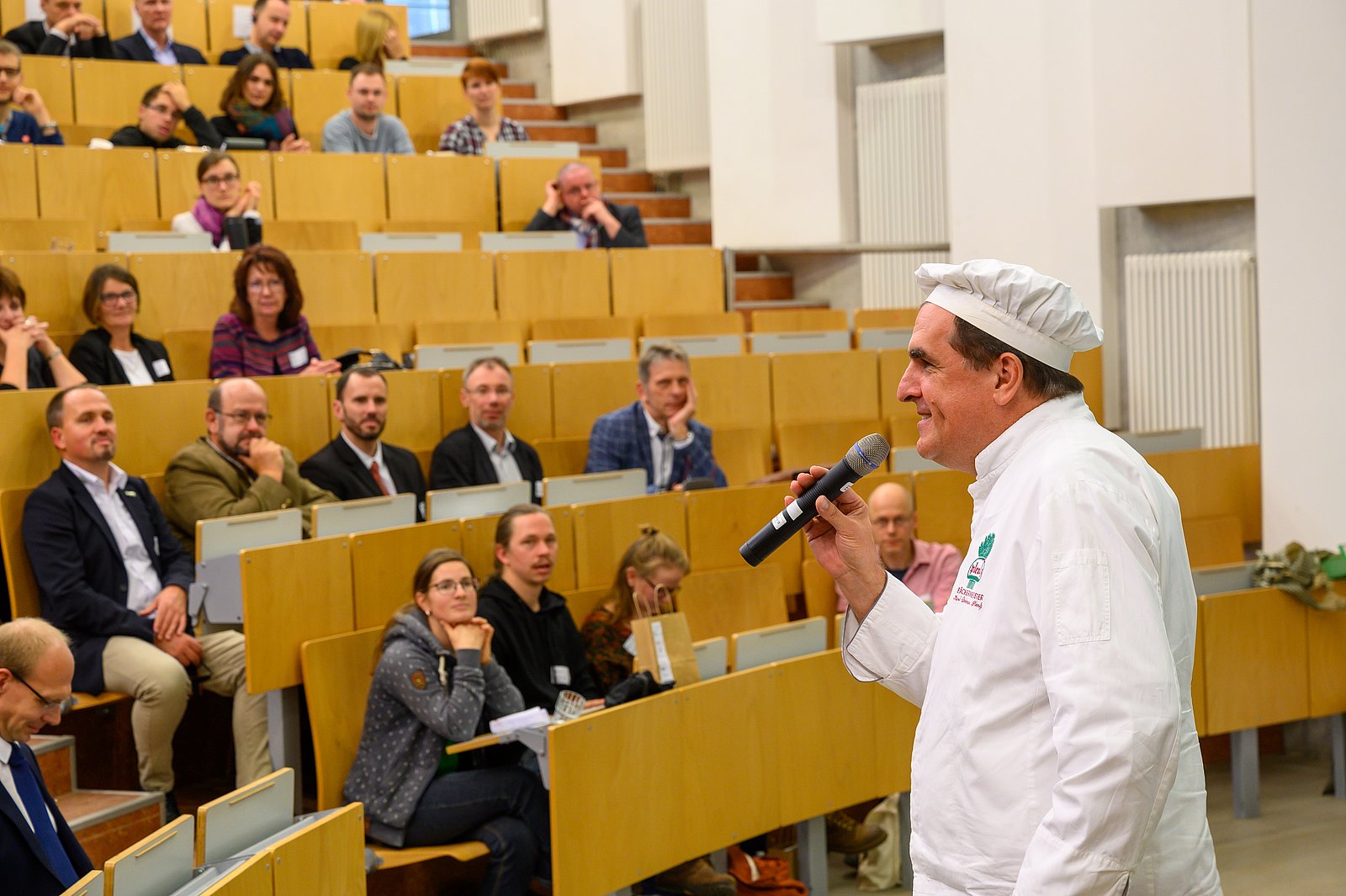Man blickt in einen Hörsaal, welcher gut gefüllt ist. Im Vordergrund steht ein Mann mit einem Mikrofon in der Hand und erzählt etwas. Seine weiße Jacke und die weiße Mütze, die er trägt, zeigen, dass er Bäcker ist.