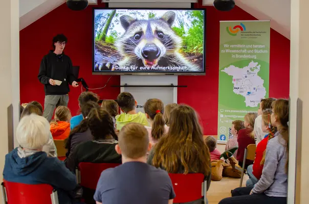 Ein Studierender präsentiert Fakten über den Waschbären vor interessierten Schüler*innen.