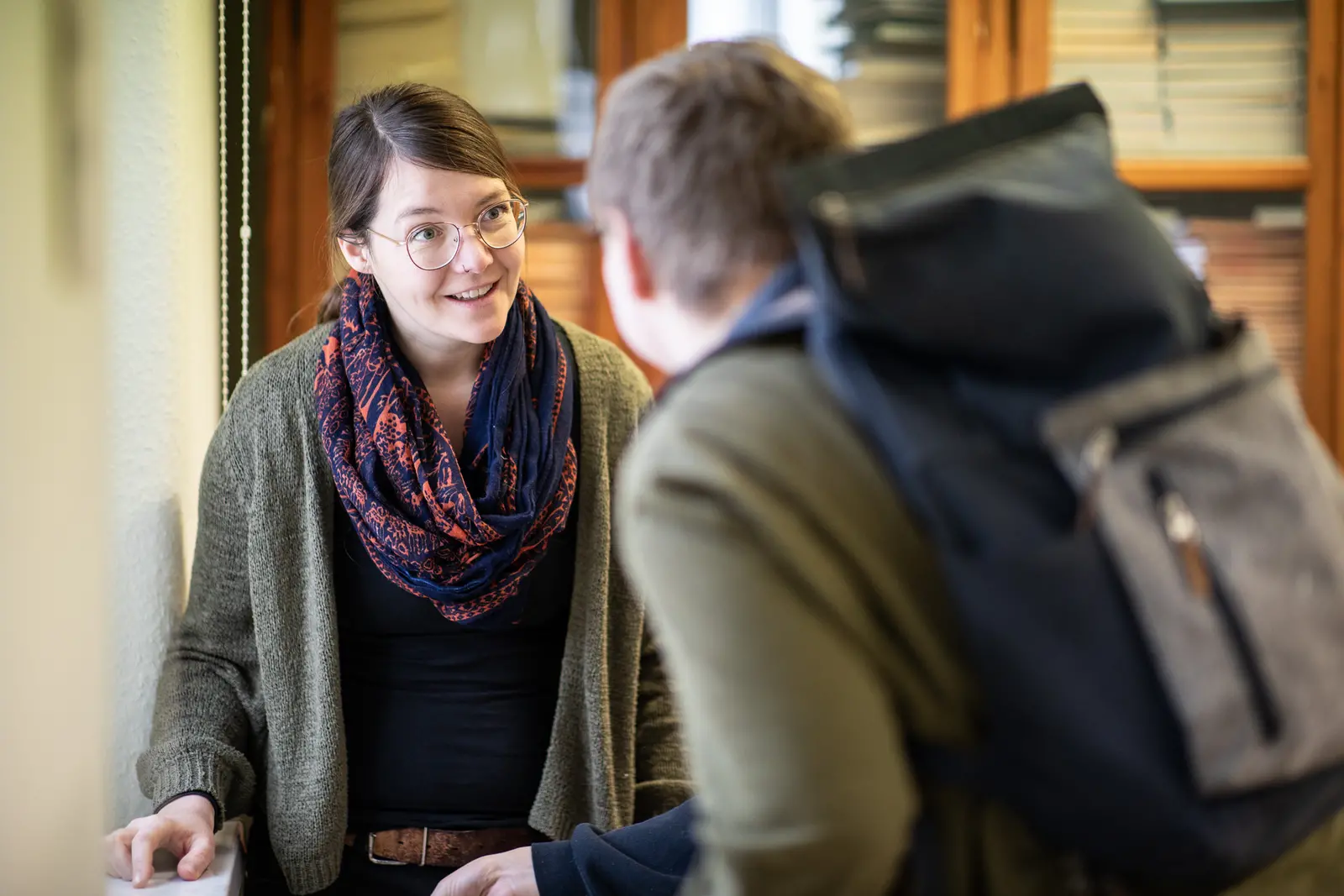 Das Bild zeigt zwei Personen in einem Gespräch während einer Veranstaltung zur Studienorientierung am Gymnasium. Eine Frau lächelt und trägt eine Brille, während sie mit einem Rucksack tragenden Jungen spricht.