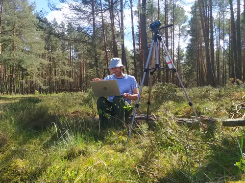 Abgebildet ist ein Kiefernwald mit moosiger Vegetation im Vordergrund. Mittig sitzt Dr. Jakob Wegener auf einem Baumstamm. Er blickt auf seinen aufgeklappten Laptop auf seinem Schoß. Auf dem Bild rechts von ihm steht eine Kamera für die Beobachtung der Bienenvölker.