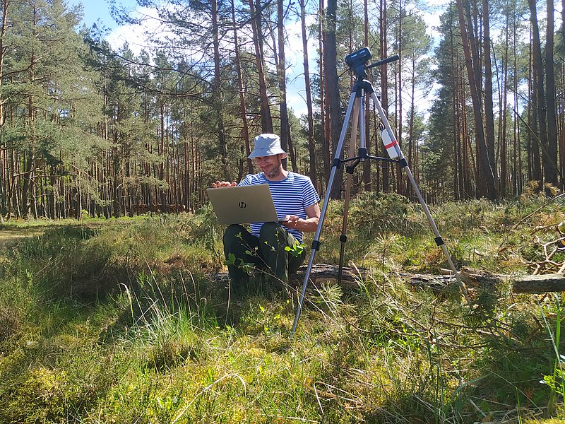 Abgebildet ist ein Kiefernwald mit moosiger Vegetation im Vordergrund. Mittig sitzt Dr. Jakob Wegener auf einem Baumstamm. Er blickt auf seinen aufgeklappten Laptop auf seinem Schoß. Auf dem Bild rechts von ihm steht eine Kamera für die Beobachtung der Bienenvölker.