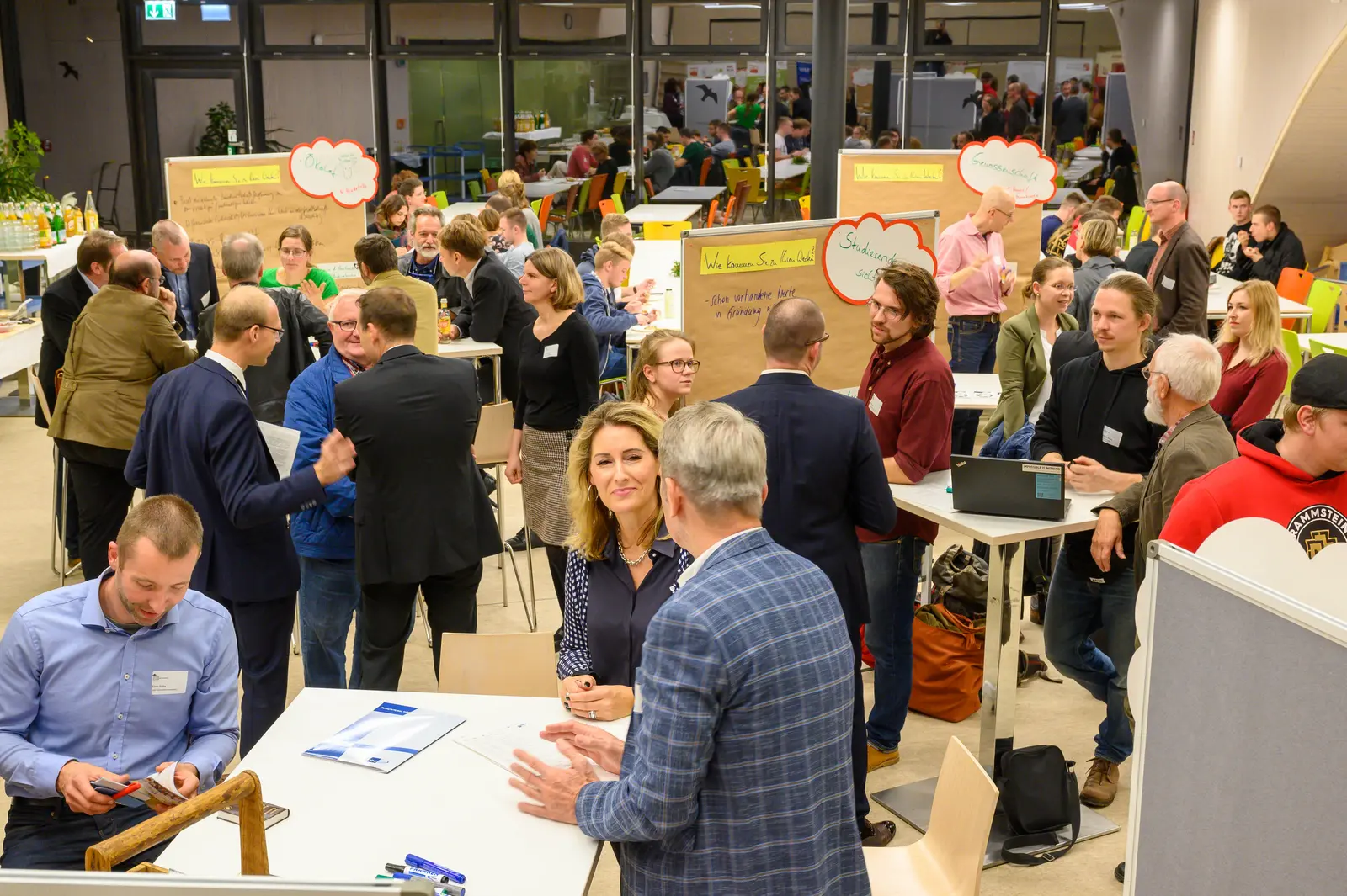 In einem sehr großen Raum stehen viele Menschen in kleinen Grüppchen an Stehtischen mit Stellwänden an den unterschiedlichste Karten pinnen und Texte geschrieben sind. Es handelt sich um einen Workshop mit Praxispartnerinnen und Praxispartnern. 