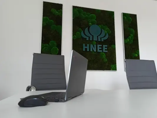 Im Vordergrund ist ein Konferenztisch zu sehen auf dem ein aufgeklappter Laptop steht. Im Hintergrund sieht man zwei graue Stuhllehnen und ein Moosbild mit HNEE-Logo an der Wand