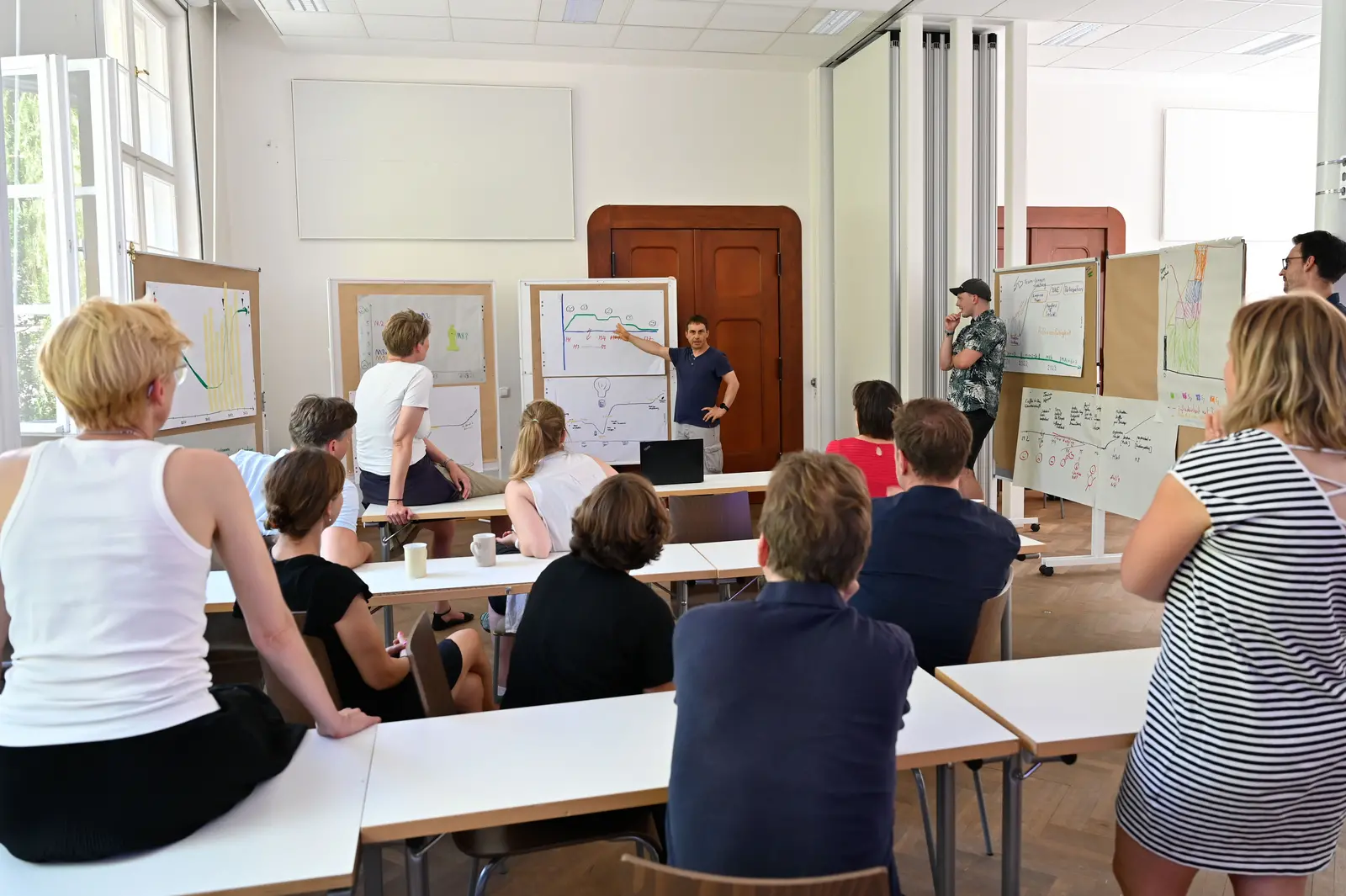 Ein Gruppe diskutiert in einem Seminarraum. Mehrere Stellwände mit während des Workshops erarbeiteten Abbildungen stehen im Halbkreis. Ein Mann erklärt gerade eine dieser Abbildungen.