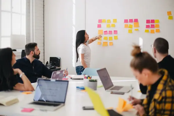 eine Gruppe von Menschen sitzt um einen Tisch während eine Frau Post-Its an der Wand arrangiert