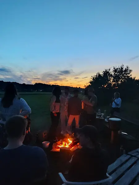 Lagerfeuer im Zentrum, verschiedene Menschen sitzen oder stehen darum und unterhalten sich. Eine Person wickelt Stockbrotteig auf. Im Hintergrund ein Sonnenuntergang