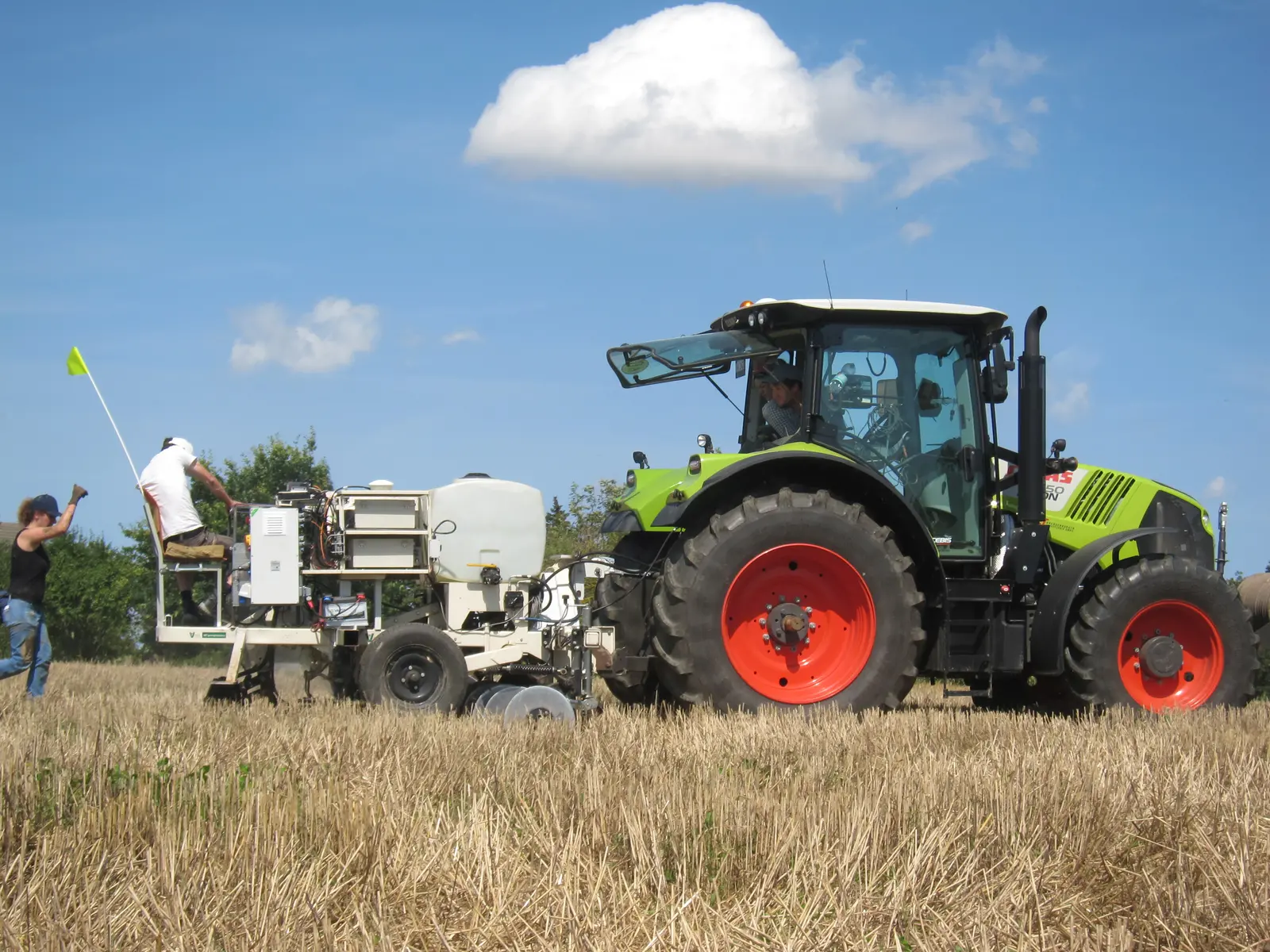 Ein Traktor zieht eine Vorrichtung zur präzisen Messung des ph-Wertes über einen Stoppelacker. Auf der Messvorrichtung sitzt ein Wissenschaftler und schaut auf den Ackerboden. Hinter der Messvorrichtung läuft eine Wissenschaftlerin. Sie schaut ebenfalls auf den Ackerboden und gibt mit dem Daumen nach oben ein positives Signal an den Wissenschaftler und den Traktorfahrer.