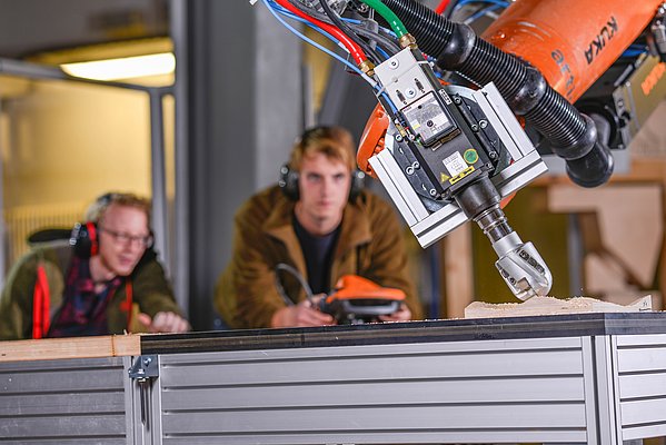 Roboterarm mit eingespanntem Fräskopf bearbeitet einen Holzprobekörper. Im Hintergrund sieht man zwei junge Wissenschaftler mit der tragbaren Robotersteuerung in der Hand.