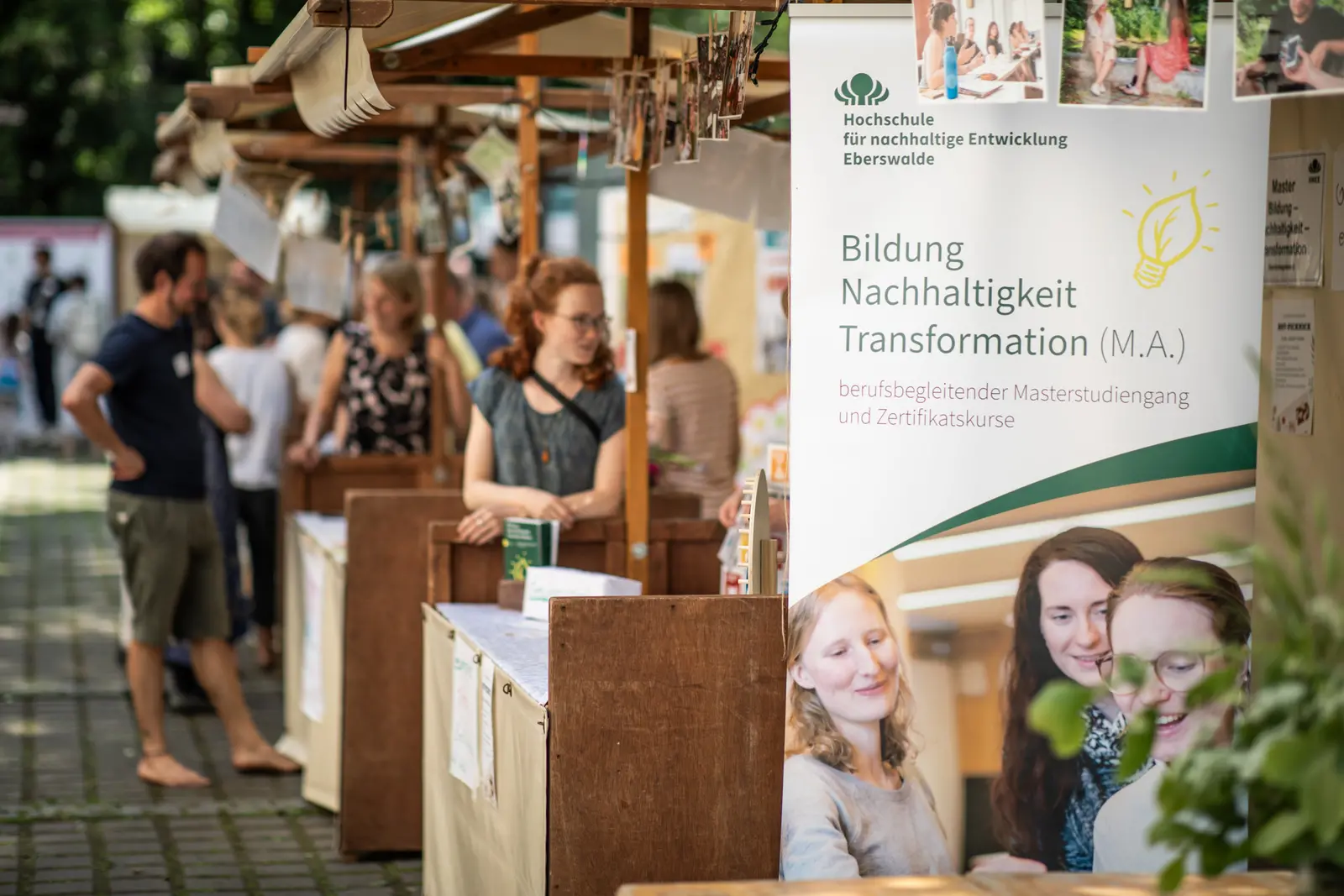 Im Vordergrund steht ein Roll-Up des Studiengangs Bildung - Nachhaltigkeit - Transformation , im Hintergrund sieht man Infostände und Personen.