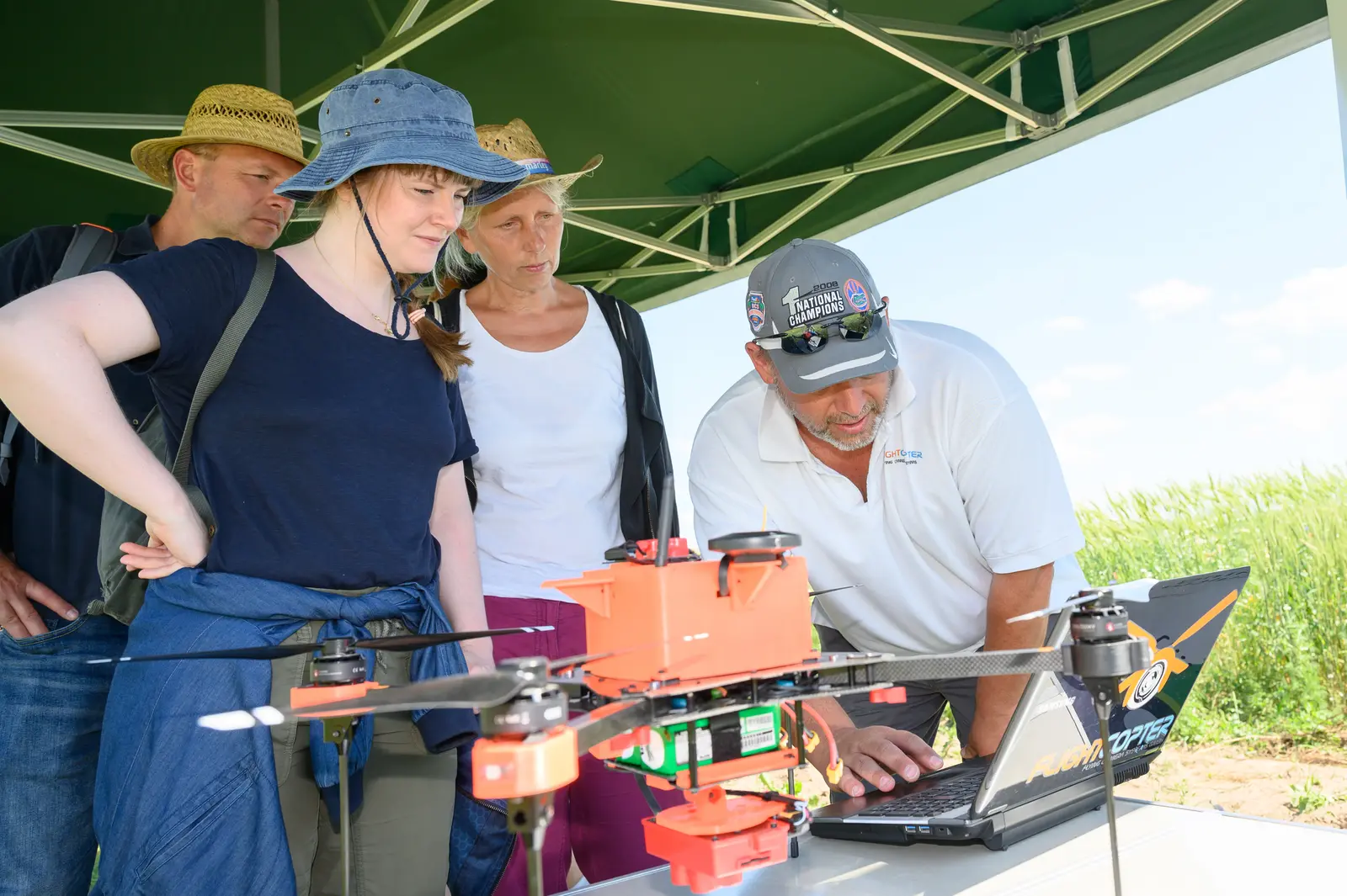 Vier Menschen stehen auf einem Feld unter einem Zelt. Vor ihnen auf einem Tisch stehen ein Laptop und eine Drohne. Die Drohne ist mit einem Sensor ausgestattet. Ein Wissenschaftler erklärt den drei Personen auf dem Laptopbildschirm den Einsatz der Drohne.