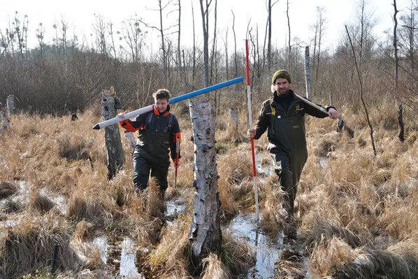 Zwei Personen laufen mit Wathosen und Werkzeugen durch ein Moor.